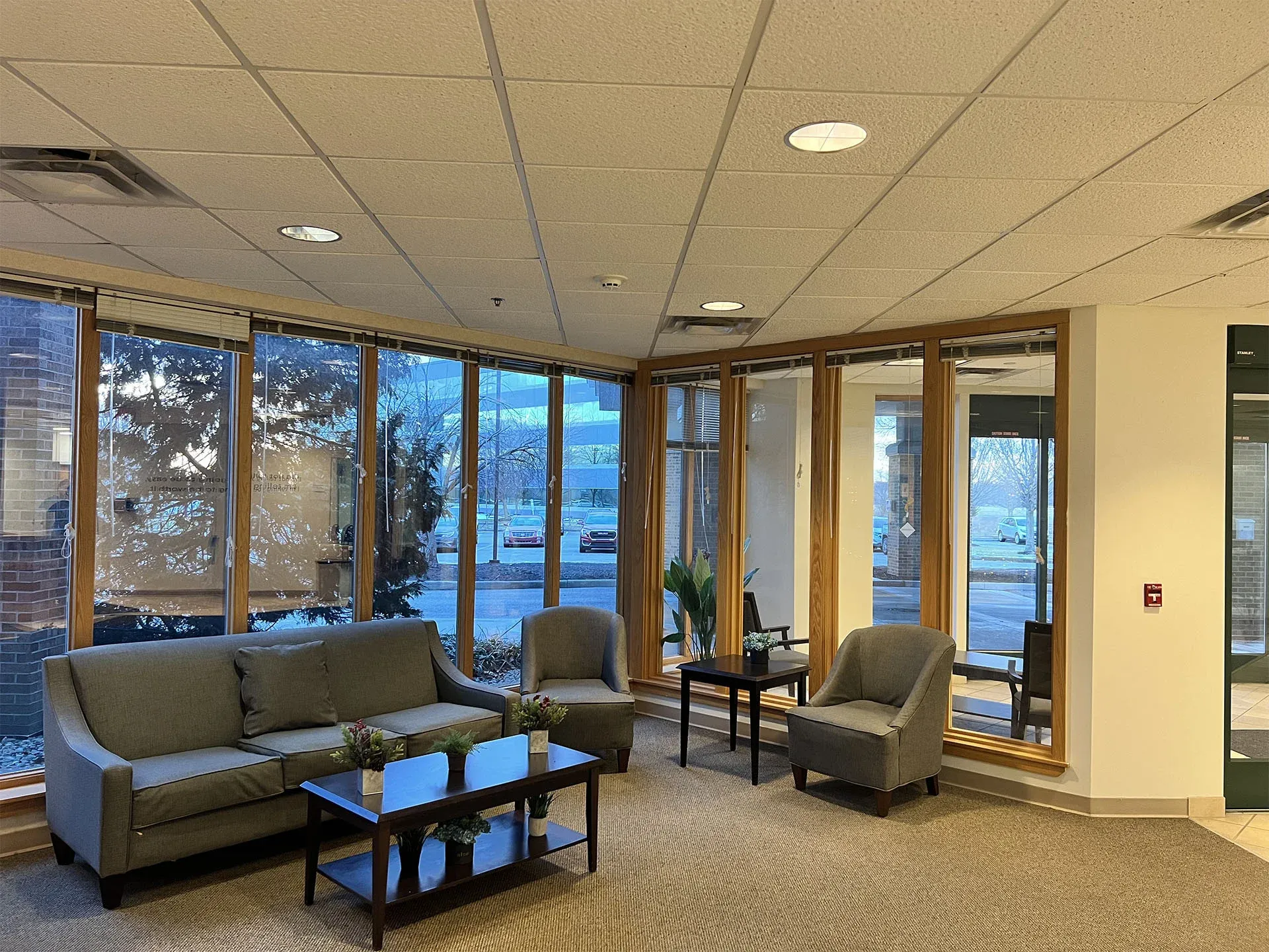 A common area at Jasper Grove — armchairs and a sofa around a low table, beside a wall of windows looking out onto trees.